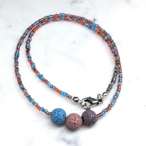 Puede incluir: Un collar de cuentas con un cierre plateado. El collar presenta una hebra de pequeñas cuentas coloridas en tonos azules, naranjas y marrones. Tres cuentas redondas de piedra de lava texturizada en azul, rosa y morado están centradas.