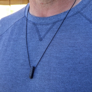 May include: A black lava rock pendant on a black cord necklace. The necklace is worn by a person wearing a blue shirt.