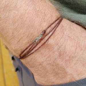 May include: A brown cord bracelet with a silver clasp. The bracelet is worn on a person's wrist.