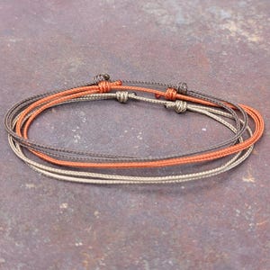 May include: A set of three thin cord bracelets in shades of dark brown, orange, and beige. Each bracelet features a sliding knot closure mechanism, allowing for adjustable sizing. The cords are smooth and round, arranged in a stacked circular formation.