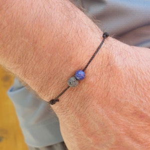 Może przedstawiać: Bransoletka ze sznurka w kolorze czarnym z koralikiem z lawy w kolorze ciemnoszarym i koralikiem z lapis lazuli w kolorze niebieskim.