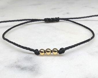 May include: A minimalist bracelet featuring three gold beads strung on a black cord. The adjustable bracelet is knotted for a secure fit. The design is simple and elegant, suitable for everyday wear.