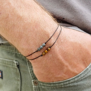 May include: Two black string bracelets, each adorned with natural stone beads. One bracelet features dark gray, porous lava beads, while the other showcases golden-brown tiger eye beads. The bracelets are displayed on a wrist.