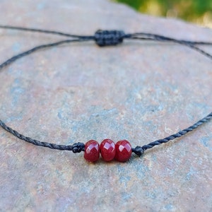 May include: A simple bracelet with three faceted, deep red beads on a black cord. The adjustable bracelet is knotted at the ends. The beads are evenly spaced, creating a minimalist design. The cord is knotted to adjust the size.