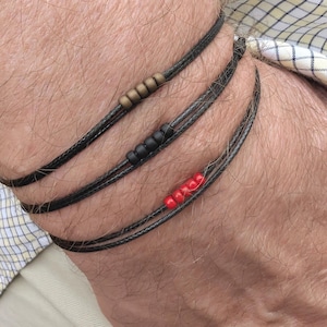 May include: Three black cord bracelets with different coloured beads. One bracelet has gold beads, another has black beads, and the third has red beads. The bracelets are wrapped around a wrist.