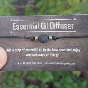 Könnte beinhalten: Ein Armband aus schwarzem Band mit einer schwarzen Lavakugel und silbernen Metallperlen. Die Karte lautet "Essential Oil Diffuser" und "Geben Sie einen Tropfen ätherisches Öl auf die Lavakugel und genießen Sie Aromatherapie unterwegs".