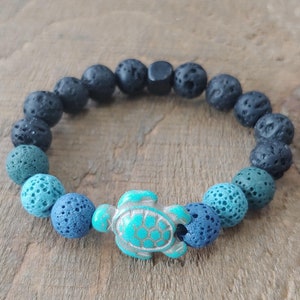 Könnte beinhalten: Ein blaues und schwarzes Lava-Armband mit einem türkisfarbenen Schildkröten-Anhänger. Das Armband besteht aus kleinen, runden Perlen und ist so gestaltet, dass es wie ein Farbverlauf aussieht, der mit Schwarz beginnt und in Blau übergeht.