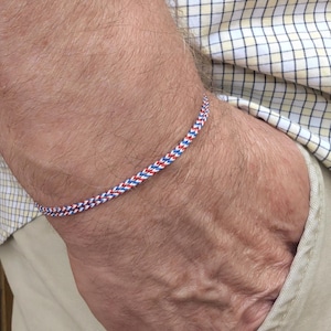 May include: A braided bracelet in red, white, and blue is shown. The bracelet is worn on a wrist. The bracelet is made of a woven material. The background includes a plaid shirt and khaki pants.