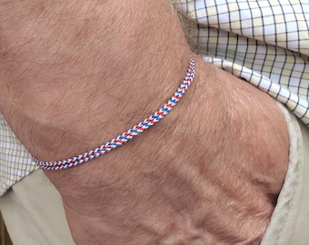 May include: A braided bracelet in red, white, and blue is shown. The bracelet is worn on a wrist. The bracelet is made of a woven material. The background includes a plaid shirt and khaki pants.