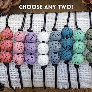 May include: A selection of lava stone bead bracelets in shades of yellow, pink, purple, white, grey, teal, green, and olive. Each bracelet has three round beads on a black cord. The text "CHOOSE ANY TWO!" is displayed at the top of the image.