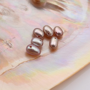 Op de afbeelding: Zes ovale, roze-paarse parels met gaten, op een parelmoer, iriserend oppervlak. De parels hebben een glanzende afwerking en reflecteren licht. De parels variëren enigszins in grootte en vorm.