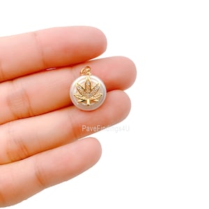 Puede incluir: Pendiente de tono dorado con un diseño de hoja de marihuana con piedras de circonita cúbica transparentes engastadas en la hoja. El pendiente está unido a una perla blanca.
