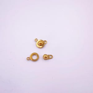 May include: Three small, gold-colored clasps are arranged on a white background. The clasps include a round ring, a circular clasp with a raised center, and a small rectangular clasp with a ball on one end.