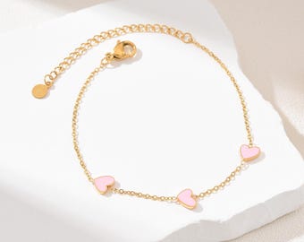 Könnte beinhalten: Ein goldfarbenes Armband mit drei rosa Herzanhängern. Das Armband hat eine verstellbare Kette und einen Karabinerverschluss. Die Herzen sind gleichmäßig verteilt. Das Armband wird auf einer weißen Oberfläche präsentiert.