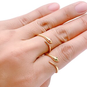 May include: Two gold snake rings with red and green gemstone eyes. The rings are worn on the middle finger of a hand.