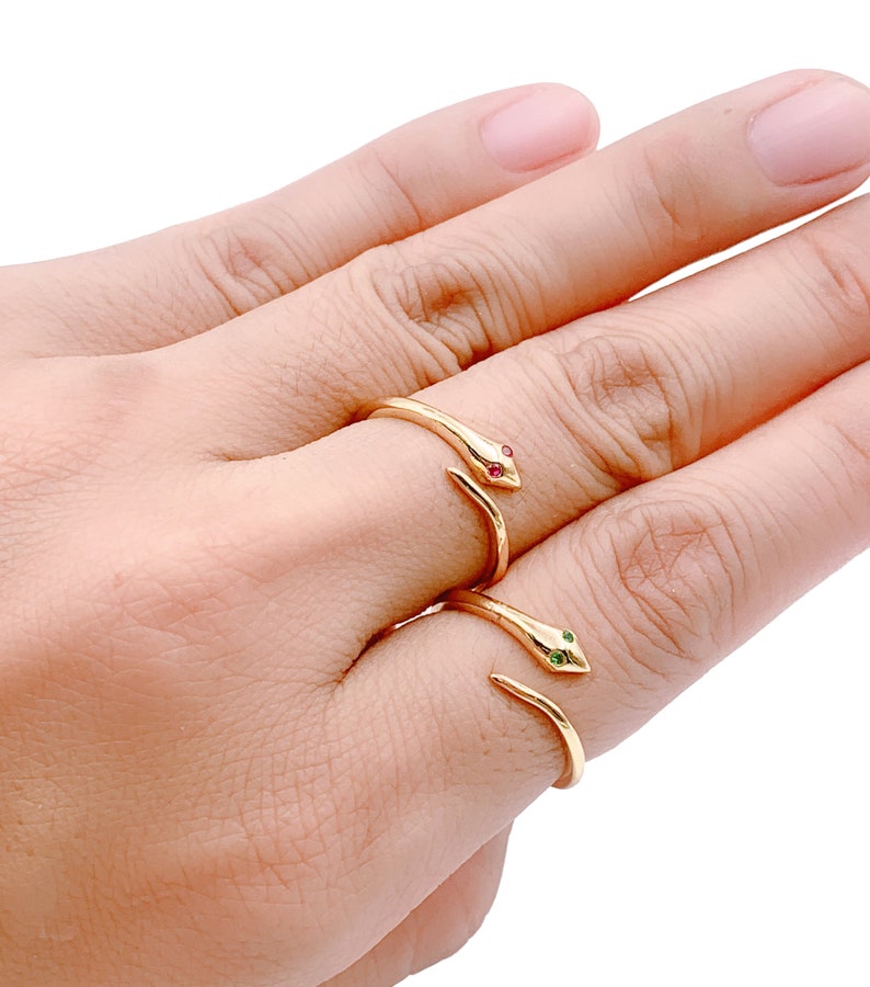 May include: Two gold snake rings with red and green gemstone eyes. The rings are worn on the middle finger of a hand.