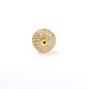 May include: A gold-toned bead with a round shape and a hole in the center. The bead is covered in small, clear rhinestones.