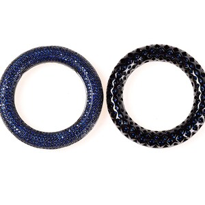 May include: Two circular bangles. One is covered in small, dark blue gemstones. The other is black with a geometric pattern and small blue gemstones. Both are set against a white background.