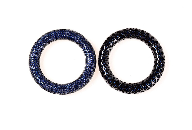 May include: Two circular bangles. One is covered in small, dark blue gemstones. The other is black with a geometric pattern and small blue gemstones. Both are set against a white background.