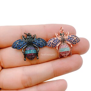Puede incluir: Dos abalorios de abeja con detalles de cristal de colores. Un abalorio es negro con cristales azules y morados, el otro es de oro rosa con cristales azules y turquesa.