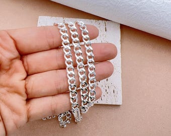 May include: A silver curb chain necklace with a polished finish. The chain is displayed on a neutral-colored surface, showcasing its links and overall design. The necklace is a classic jewelry piece.