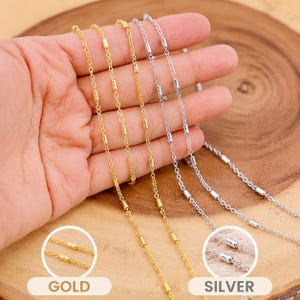 May include: Close-up of gold and silver necklaces with a delicate chain design. The necklaces feature small, cylindrical beads spaced along the chain. The word "GOLD" is visible in a circle, and the word "SILVER" is visible in another circle.