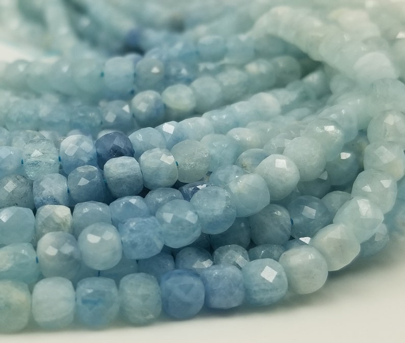 May include: Close-up of aquamarine gemstone beads. The beads are a light blue color with faceted surfaces, creating a sparkling effect. The beads are strung together, suggesting they are for jewelry making or crafting.