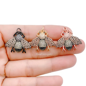 May include: Three bee-shaped pendants in black, gold, and rose gold. Each pendant features a body with black and gold stripes, wings covered in small, sparkling stones, and a loop for attaching to jewelry. The pendants are held in a hand.