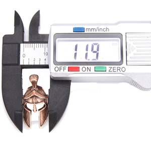 May include: A bronze-colored Spartan helmet pendant is measured with a digital caliper. The helmet is approximately 0.47 inches in length. The caliper displays a measurement of 11.9 mm. The background is white.