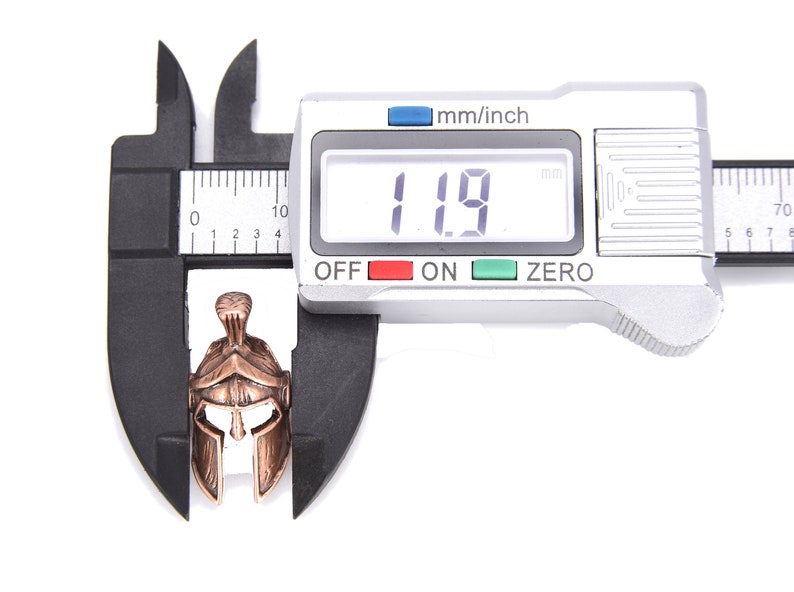 May include: A bronze-colored Spartan helmet pendant is measured with a digital caliper. The helmet is approximately 0.47 inches in length. The caliper displays a measurement of 11.9 mm. The background is white.