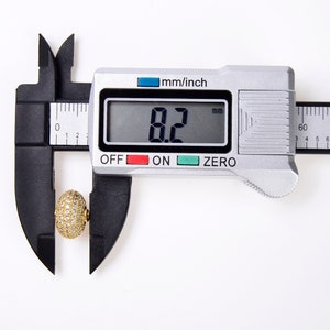May include: A close-up of a digital caliper measuring a gold-colored, round object with small white stones. The caliper reads 8.2 mm.