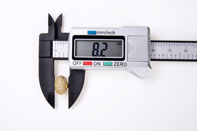 May include: A close-up of a digital caliper measuring a gold-colored, round object with small white stones. The caliper reads 8.2 mm.