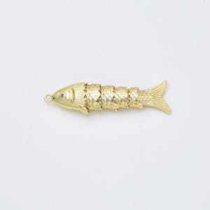 May include: A gold-colored fish pendant with a textured, segmented body and a small loop at the head. The fish has a detailed fin and tail design, and a small gem is set in the eye. The pendant is approximately 3 inches long.