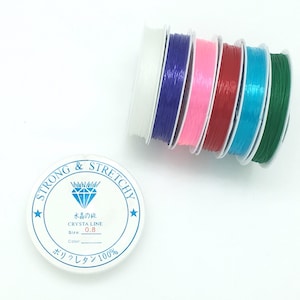 May include: A spool of clear elastic string with a label that reads "STRONG & STRETCHY" and "CRYSTA LINE 0.8". The spool is white with a blue diamond and stars. The elastic string is available in six colors: white, purple, pink, red, blue, and green.
