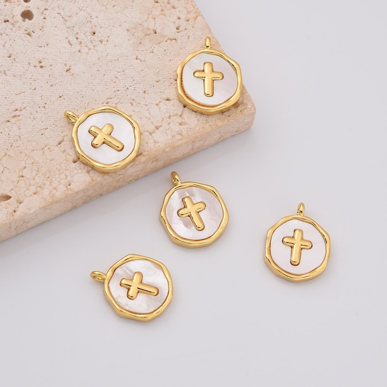 Op de afbeelding: Vijf goudkleurige metalen bedels met een witte schelp inlay en een gouden kruisontwerp.