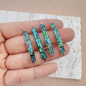May include: Four rectangular bar pendants with iridescent abalone shell inlays. The bars are framed in gold and silver metal. The abalone shell displays shades of green, blue, and purple. The pendants are displayed on a neutral background.