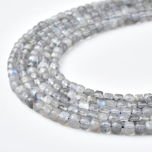 May include: Strands of faceted, gray labradorite beads. The beads are square-shaped and strung together, displaying a subtle iridescence. The beads are on a white background.