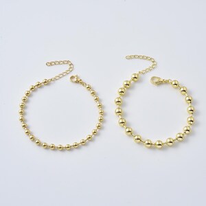 May include: Two gold-colored beaded bracelets with adjustable chain closures. The bracelets feature small, evenly spaced spherical beads. The clasps and chains are also gold-toned, creating a cohesive and elegant look. The jewelry is displayed on a white surface.