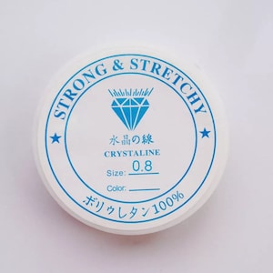 May include: A white round container with blue text and a diamond logo. The text reads "STRONG & STRETCHY", "水晶の線", "CRYSTALINE", "Size:", "Color:", "0.8", and "ボリウレタン100%".