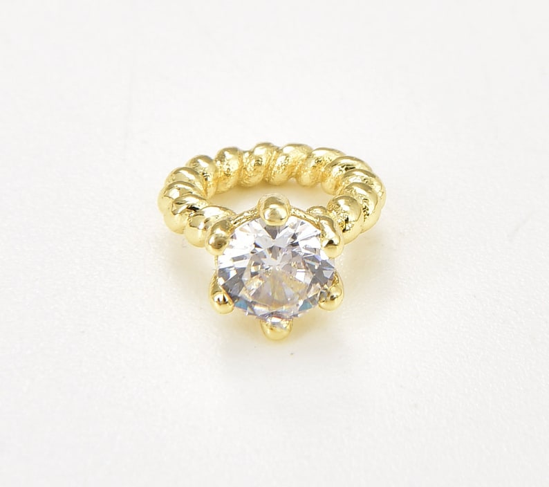 May include: A gold ring featuring a large, clear, round gemstone. The ring band has a twisted rope design. The gemstone is held by six prongs. The ring is set against a white background.