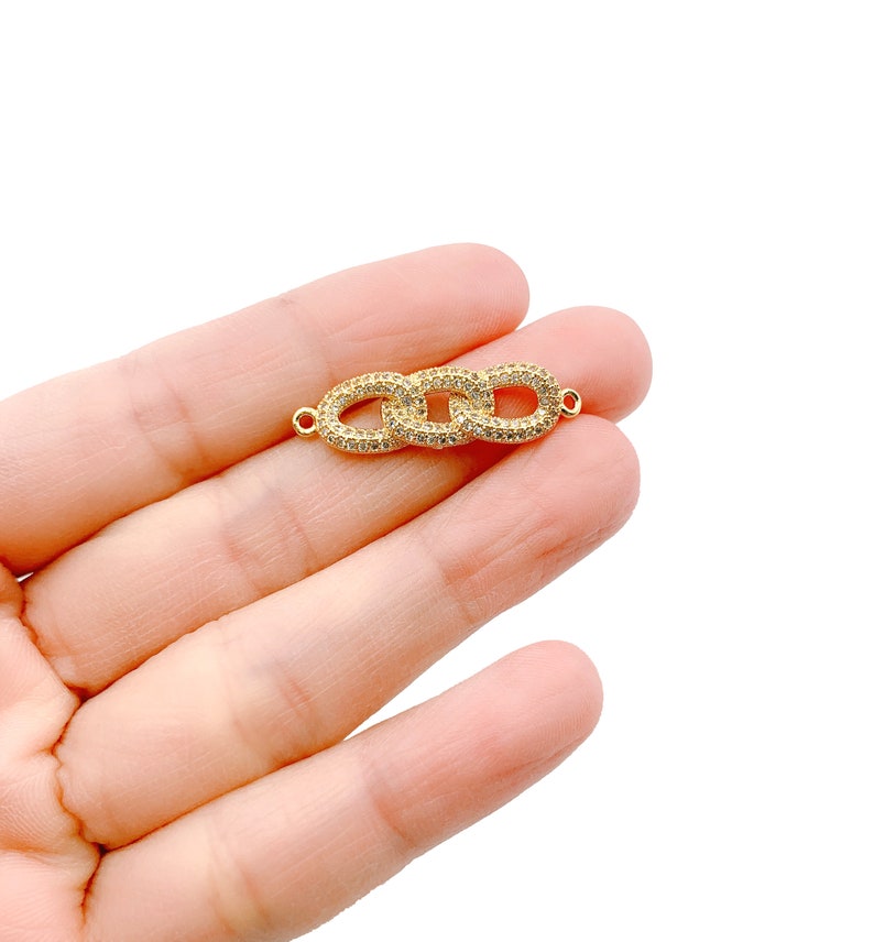 May include: A gold-colored jewelry connector with three oval links, each encrusted with small, clear crystals. The connector is held in a person's hand against a white background. The connector has small loops on each end.