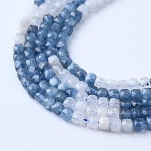 May include: Close-up of strands of faceted beads in shades of blue and white. The beads are strung together, creating a textured and layered effect. The beads have a geometric shape and a polished finish, reflecting light.