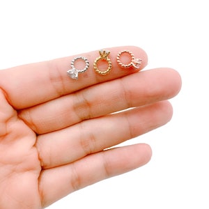 May include: Three small ring-shaped earrings in silver, gold, and rose gold. Each earring features a twisted band and a clear gemstone. The earrings are displayed on a light background.