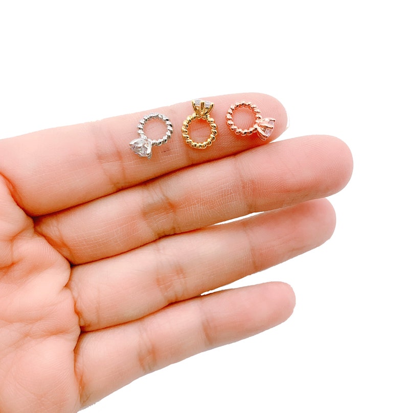 May include: Three small ring-shaped earrings in silver, gold, and rose gold. Each earring features a twisted band and a clear gemstone. The earrings are displayed on a light background.