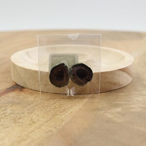 May include: Two halves of a dark olive are displayed in a clear plastic square. The olive halves are positioned in front of a small, round wooden dish. The background is a light brown wooden surface.