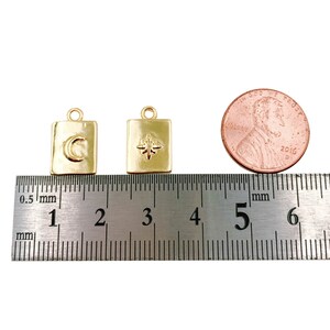 May include: Two gold-toned charms, each with a square shape and a different design. The charm on the left has a crescent moon design, while the charm on the right has a star design. The charms are measured against a ruler, showing they are approximately 1.5 cm in size.