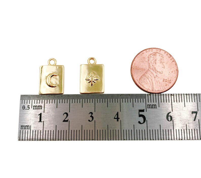 May include: Two gold-toned charms, each with a square shape and a different design. The charm on the left has a crescent moon design, while the charm on the right has a star design. The charms are measured against a ruler, showing they are approximately 1.5 cm in size.