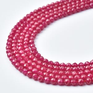 May include: Close-up of several strands of faceted, deep red gemstone beads. The beads are small, round, and evenly sized, creating a textured, flowing appearance. The background is plain white, highlighting the vibrant color and detail of the gemstones.