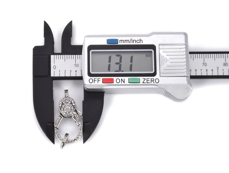 May include: A silver-toned clasp with clear cubic zirconia stones, measured at 13.1 mm using a digital caliper.