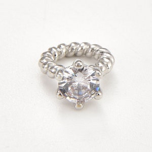 May include: A silver ring featuring a large, clear, faceted gemstone. The ring band has a twisted rope design. The gemstone is round with multiple facets, reflecting light. The ring is set against a white background.